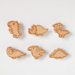 Puede incluir: Seis recortes de dinosaurios de madera en varias formas, incluyendo un T-Rex, Triceratops y Estegosaurio. Cada dinosaurio es marrón claro con detalles grabados, sobre un fondo blanco. Ideales para manualidades infantiles o juguetes educativos.