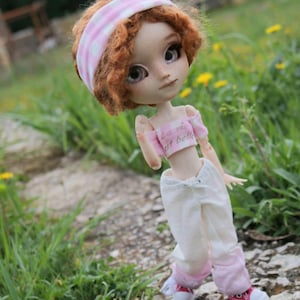 Può includere: Una bambola con capelli rossi ricci che indossa una fascia rosa e bianca, un top corto rosa con la scritta "La vie est belle" e pantaloni bianchi con rifiniture rosa. La bambola indossa scarpe da ginnastica rosse e si trova su un sentiero.