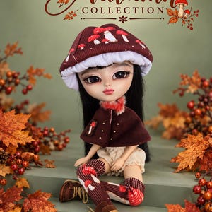 Pode incluir: Uma boneca com cabelos pretos longos usando um chapéu marrom escuro com estampa de cogumelos, uma capa marrom com pequenos tachas douradas, shorts cor de creme e meias altas combinando com estampa de cogumelos e botas marrons. O texto Autumn COLLECTION é visível.