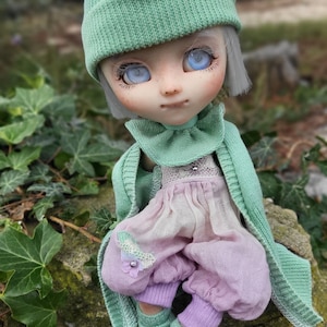 Könnte beinhalten: Eine Puppe mit weißen Haaren, die einen grünen Strickhut, ein rosa-grünes Kleid und grüne Strickgamaschen trägt. Die Puppe sitzt auf einem mit Moos bewachsenen Stein.