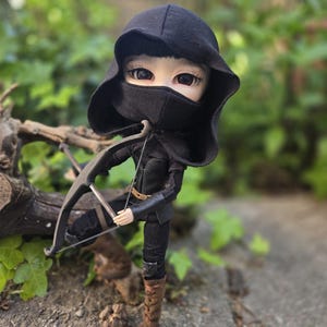 May include: A doll dressed in a black hooded outfit and mask, holding a bow and arrow. The doll wears a black outfit with brown boots. The background is a blurred outdoor scene with green foliage and a tree trunk.