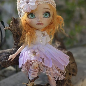 Könnte beinhalten: Eine Porzellanpuppe mit langen, lockigen, roten Haaren, die einen weißen Spitzenhut mit einer Blume, ein weißes und lavendelfarbenes Kleid mit Spitzenbesatz und braune Stiefel trägt.