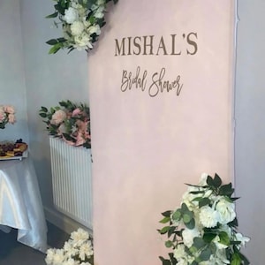Può includere: Un alto fondale ad arco color rosa pallido con il testo scuro "MISHAL'S" in un carattere serif in grassetto e "Bridal Shower" in corsivo. L'espositore è decorato con grappoli di fiori bianchi e foglie verdi nella parte superiore e inferiore.