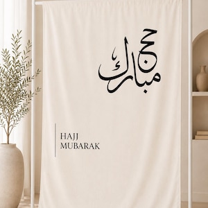 Puede incluir: Un telón de fondo de tela color crema colgado en un soporte de metal blanco. La tela presenta caligrafía árabe negra en la parte superior y el texto en inglés "HAJJ MUBARAK" impreso en una fuente negra sin remates, junto a una línea vertical.