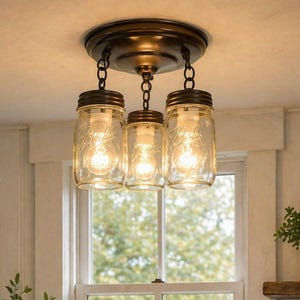 Peut inclure: Un luminaire de plafond de style rustique avec trois abat-jours en verre en forme de bocaux Mason. Le luminaire a une finition bronze foncé et est suspendu à des chaînes. Les bocaux contiennent des ampoules chaudes et lumineuses.