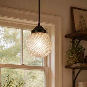 May include: A pendant light fixture with a textured, spherical glass shade hangs in front of a window. The light has a dark metal base and a warm glow. The kitchen setting includes a sink, shelves, and plants.