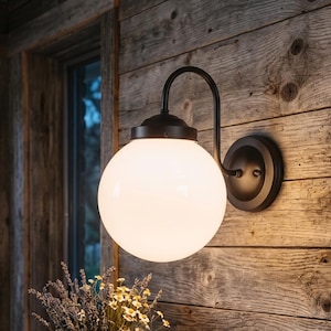 May include: A black wall-mounted globe light with a white glass shade. The light is fixed to a wooden wall. Below, a wooden table holds a book, glasses, a mug, and a small flower pot. The room has a rustic, farmhouse style.