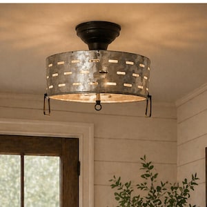May include: A rustic ceiling light fixture with a galvanized metal shade. The shade has a cylindrical shape with evenly spaced rectangular cutouts. The fixture is mounted on a dark brown base and hangs from the ceiling. The light is on, illuminating the room.
