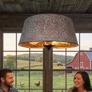 Puede incluir: Una familia disfruta de una comida en una mesa de madera en una cocina r&uacute;stica. Una gran pantalla de l&aacute;mpara estampada cuelga sobre la mesa. La ventana ofrece una vista de un granero rojo y campos verdes. Ollas y sartenes de cobre est&aacute;n en los estantes.