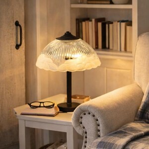 May include: Cozy living room scene with a beige armchair, plaid throw blanket, and decorative pillow. A lit lamp with a floral shade sits on a side table with books and glasses. A framed painting and bookshelf complete the warm, inviting space.
