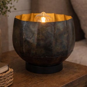 May include: A decorative table lamp with a dark, textured exterior and a gold-colored interior. The lamp has a round shape and sits on a black base. A lit bulb is visible inside. The lamp is on a wooden nightstand.