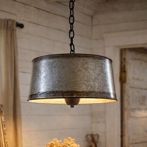 May include: A metal pendant light with a galvanised finish hangs from a chain. A wooden table below displays a vintage lantern, dried flowers in a glass jar, and woven baskets. The room has a farmhouse aesthetic.