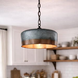 May include: A rustic kitchen pendant light with a galvanized metal shade and copper trim hangs above a marble countertop. A wooden tray holds a vase of white flowers, a pepper grinder, and lemons. Woven stools sit below.