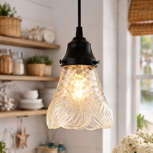May include: A pendant light fixture with a black metal base and a clear, textured glass shade. The light is on, illuminating the intricate patterns within the shade. The kitchen setting includes wooden shelves, white cabinets, and a coastal theme.
