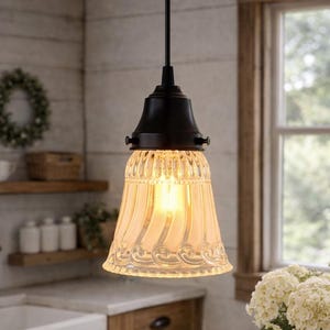 May include: A vintage-style pendant light with a clear, fluted glass shade and a dark bronze-colored fixture. The light hangs above a wooden table in a kitchen setting, with a farmhouse aesthetic. The shade has a decorative swirl pattern.