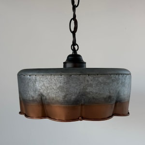 May include: A hanging pendant light with a galvanized metal shade and a copper-colored bottom edge. The light fixture is suspended from a black chain and canopy. The shade has a unique, scalloped design.