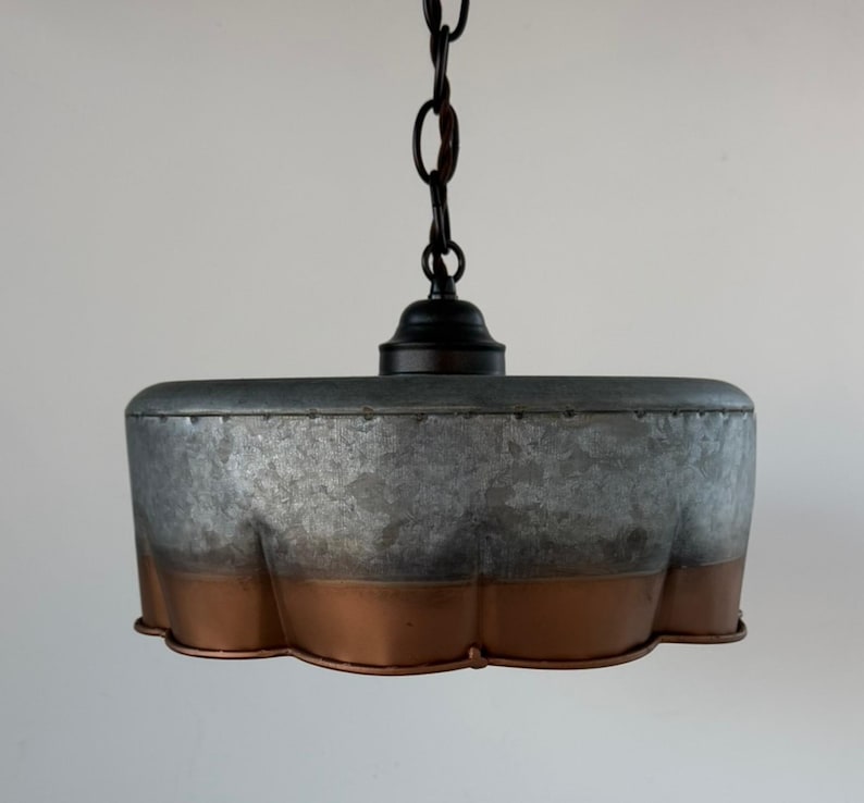 May include: A hanging pendant light with a galvanized metal shade and a copper-colored bottom edge. The light fixture is suspended from a black chain and canopy. The shade has a unique, scalloped design.