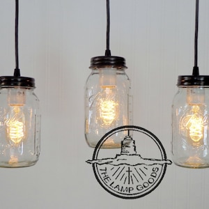 Puede incluir: Tres tarros de cristal con tapas negras cuelgan de cordones negros. Cada tarro tiene una bombilla en su interior y está encendido. Los tarros cuelgan de una pared blanca. El logotipo de The Lamp Goods es visible en la parte inferior central de la imagen.