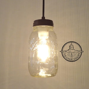 May include: A clear glass mason jar pendant light fixture with a brown metal top and a light bulb inside. The jar has the words "Ball" and "Mason" printed on it. The light is hanging from a brown cord.