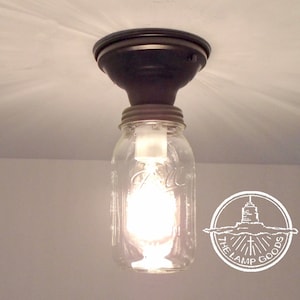 May include: A glass jar ceiling light fixture with a black metal canopy and a clear light bulb. The jar has a metal lid and is hanging from the ceiling. The light is on and the bulb is glowing. The logo "The Lamp Goods" is visible in the lower right corner of the image.