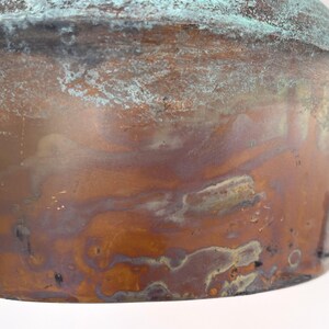 May include: Close-up of a copper object with a weathered, aged appearance. The surface displays a mix of brown, green, and blue-green hues, with visible oxidation and texture. The object's shape is rounded, suggesting it could be a bowl or a similar vessel.