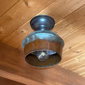 May include: A rustic ceiling light fixture with a copper-colored shade and a dark metal base. The shade has a turquoise patina, and a clear lightbulb is visible inside. The fixture is mounted on a wooden ceiling.