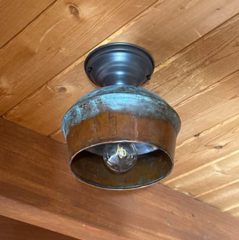 May include: A rustic ceiling light fixture with a copper-colored shade and a dark metal base. The shade has a turquoise patina, and a clear lightbulb is visible inside. The fixture is mounted on a wooden ceiling.