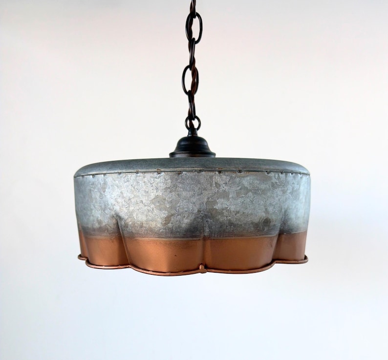 May include: A hanging pendant light with a galvanized metal shade and a copper-colored bottom edge. The light fixture is suspended from a dark chain and has a black fixture cap. The shade has a unique, scalloped edge.