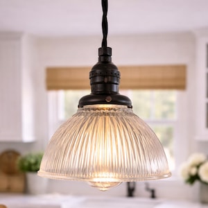 May include: A vintage-style pendant light fixture with a clear, ribbed glass shade and a dark bronze-colored metal base. The light hangs from a black cord, illuminating a kitchen setting. The shade is approximately 8 inches in diameter.