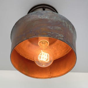 May include: A vintage-style ceiling light fixture with a copper-colored shade and a visible Edison bulb. The fixture has a circular design with a distressed finish, and the bulb is lit, casting a warm glow.