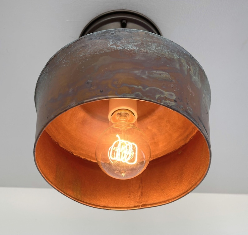 May include: A vintage-style ceiling light fixture with a copper-colored shade and a visible Edison bulb. The fixture has a circular design with a distressed finish, and the bulb is lit, casting a warm glow.