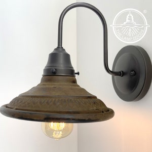May include: A vintage-style wall sconce with a dark bronze finish. The lamp features a decorative, aged-looking shade and a curved arm. The light bulb is visible, emitting a warm glow. The fixture is mounted on a round base, perfect for interior lighting.