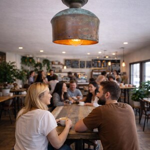 May include: A copper-colored pendant light fixture hangs above a wooden table in a cafe. The light has a cylindrical shade and a warm, inviting glow. People are seated around the table, enjoying coffee and conversation.