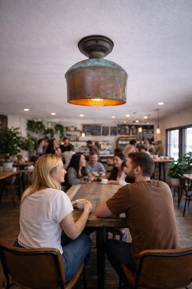 May include: A copper-colored pendant light fixture hangs above a wooden table in a cafe. The light has a cylindrical shade and a warm, inviting glow. People are seated around the table, enjoying coffee and conversation.