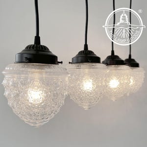 May include: Four pendant lights with clear, textured glass shades and black hardware. The shades have a unique, bubbly texture, and the lights are suspended from black cords. The Lamp Goods logo is visible in the upper right corner.