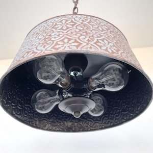 Puede incluir: Una l&aacute;mpara colgante r&uacute;stica con una pantalla estampada de color cobre y un interior negro. La l&aacute;mpara tiene cinco bombillas Edison de estilo vintage. La pantalla est&aacute; suspendida de una cadena.