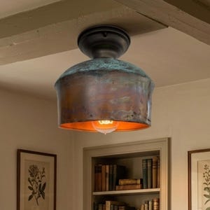 May include: A ceiling light with a copper-toned shade and a black base. The shade has a weathered appearance with a green patina. The light bulb is visible, emitting a warm, orange glow. The fixture is mounted on a white ceiling.