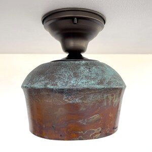 May include: A ceiling-mounted light fixture with a copper-colored shade and a bronze-colored mounting plate. The shade has a rounded top with a turquoise patina and a flared, cylindrical base. The fixture is designed for indoor use.