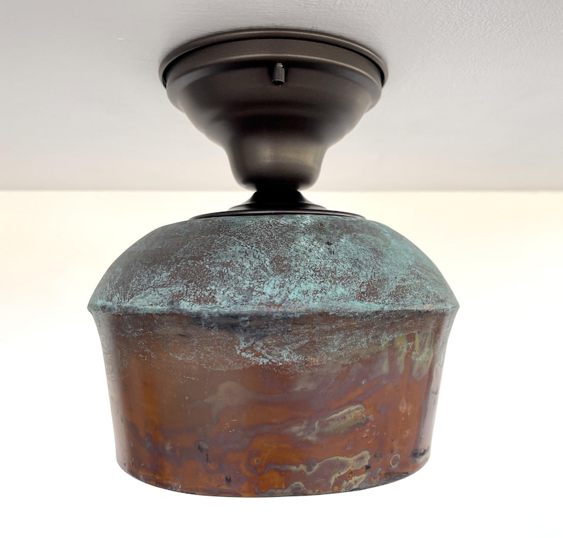 May include: A ceiling-mounted light fixture with a copper-colored shade and a bronze-colored mounting plate. The shade has a rounded top with a turquoise patina and a flared, cylindrical base. The fixture is designed for indoor use.
