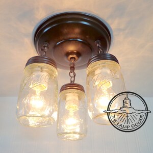 May include: A rustic ceiling light fixture featuring three clear glass Ball mason jars suspended from a bronze-colored metal base. Each jar houses a lit bulb, creating a warm, inviting glow. The fixture is designed for indoor use.