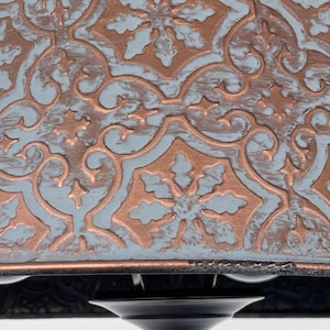 Puede incluir: Primer plano de una l&aacute;mpara de techo decorativa. La l&aacute;mpara presenta un patr&oacute;n detallado en relieve en cobre y azul claro. El dise&ntilde;o incluye motivos florales y de volutas. La base de la l&aacute;mpara es negra.