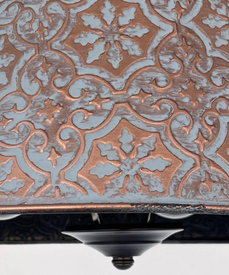 Puede incluir: Primer plano de una l&aacute;mpara de techo decorativa. La l&aacute;mpara presenta un patr&oacute;n detallado en relieve en cobre y azul claro. El dise&ntilde;o incluye motivos florales y de volutas. La base de la l&aacute;mpara es negra.
