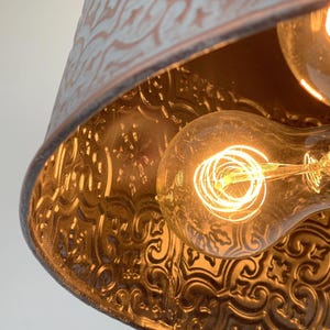 Puede incluir: Primer plano de una l&aacute;mpara colgante de estilo vintage. La l&aacute;mpara tiene una pantalla de metal desgastado, azul claro y marr&oacute;n, con un patr&oacute;n floral en relieve. Se ve una bombilla Edison c&aacute;lida y brillante en el interior.