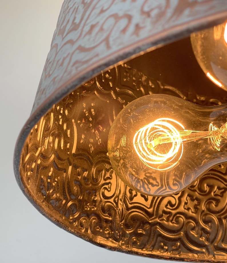 Puede incluir: Primer plano de una l&aacute;mpara colgante de estilo vintage. La l&aacute;mpara tiene una pantalla de metal desgastado, azul claro y marr&oacute;n, con un patr&oacute;n floral en relieve. Se ve una bombilla Edison c&aacute;lida y brillante en el interior.