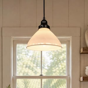 May include: A pendant light fixture with a white, textured shade and a black metal base. The light hangs from a black cord and is positioned in front of a window. The shade has a conical shape and emits a warm glow.