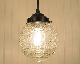 May include: A clear glass pendant light fixture with a textured, diamond-patterned globe shade. The fixture has a dark metal cap and stem. The light is on, illuminating the patterned glass.