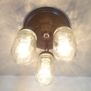 May include: A three-light ceiling fixture featuring clear glass mason jars as shades. The fixture has a bronze-colored base and metal accents. Each jar is illuminated by a bright light bulb, creating a rustic, farmhouse aesthetic.
