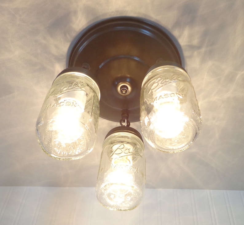 May include: A three-light ceiling fixture featuring clear glass mason jars as shades. The fixture has a bronze-colored base and metal accents. Each jar is illuminated by a bright light bulb, creating a rustic, farmhouse aesthetic.