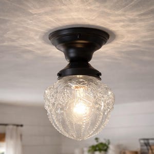 May include: A vintage-style ceiling light fixture with a clear, patterned glass shade and a dark bronze-colored base. The glass shade has a detailed floral design. The light is on, emitting a warm glow. The fixture is mounted on a white ceiling.