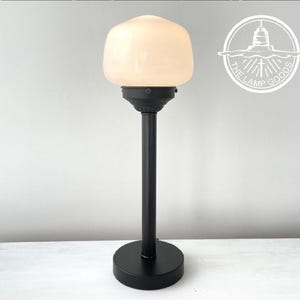 May include: Black table lamp with a white glass shade. The lamp has a tall, slender base and a round, flat base. The lamp is on a white surface. The lamp is from The Lamp Goods.